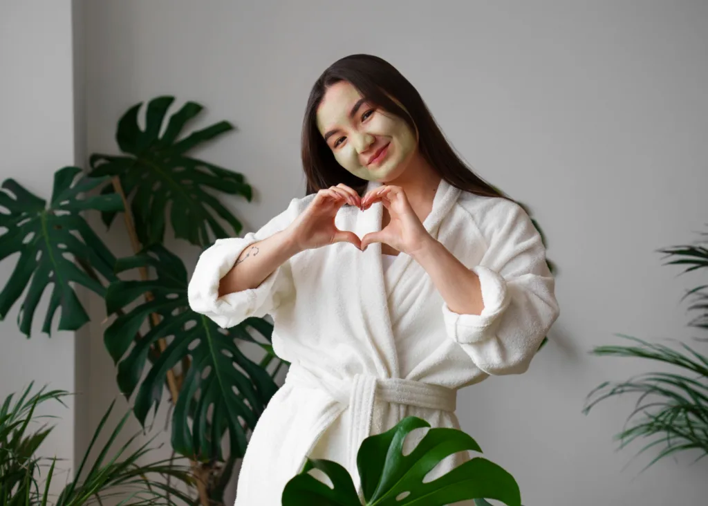 jovem oriental de roupão, usando uma creme facial, fazendo um coração com as mãos.