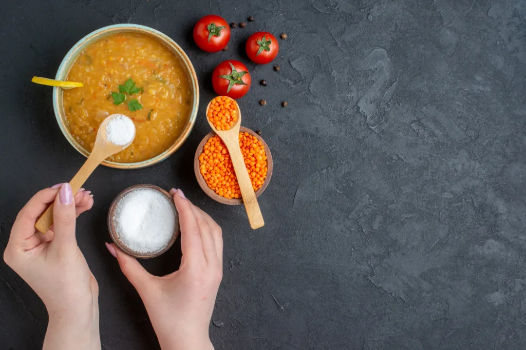 Tigela com uma sopa, sendo temperada com sal. alguns ingredientes ao redor da panela.