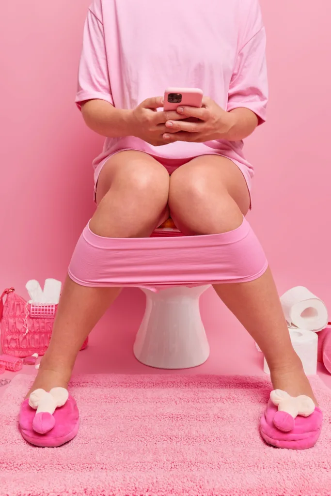 mulher de roupa cor de rosa, sentada num vazo sanitário de um banheiro de paredes cor de rosa,