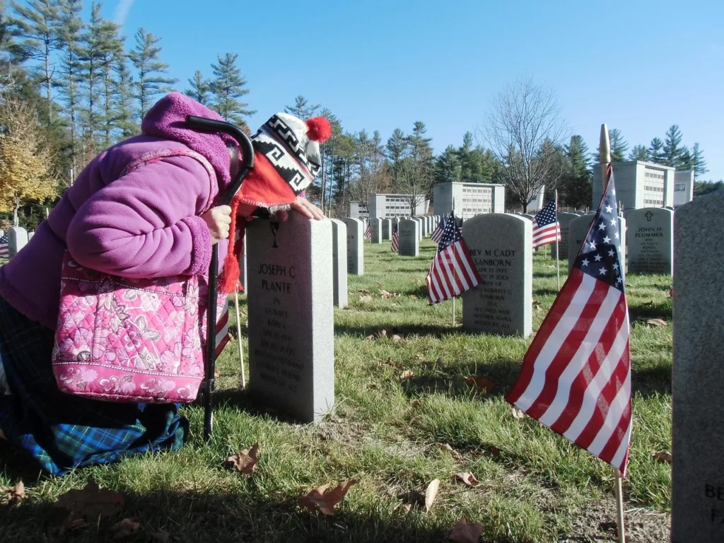 uma senhora em trajes de frio rezando em uma lapide dee um cemitério de veteranos com a bandeira americana.