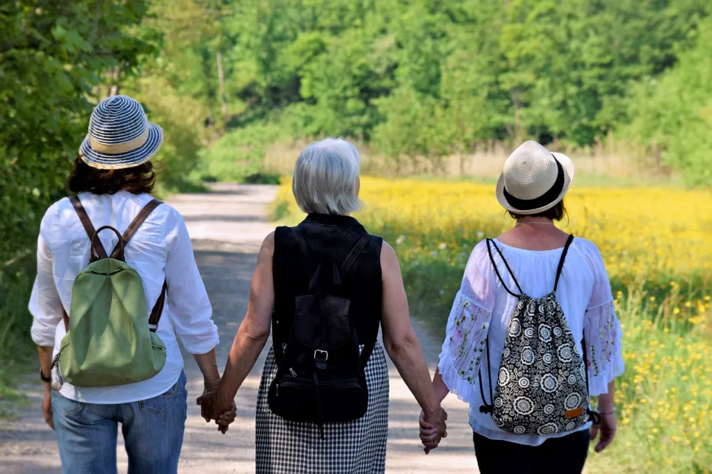 três amigas de meia idade, vistas de costas, de mãos dadas, caminhando em uma trilha de floresta.
