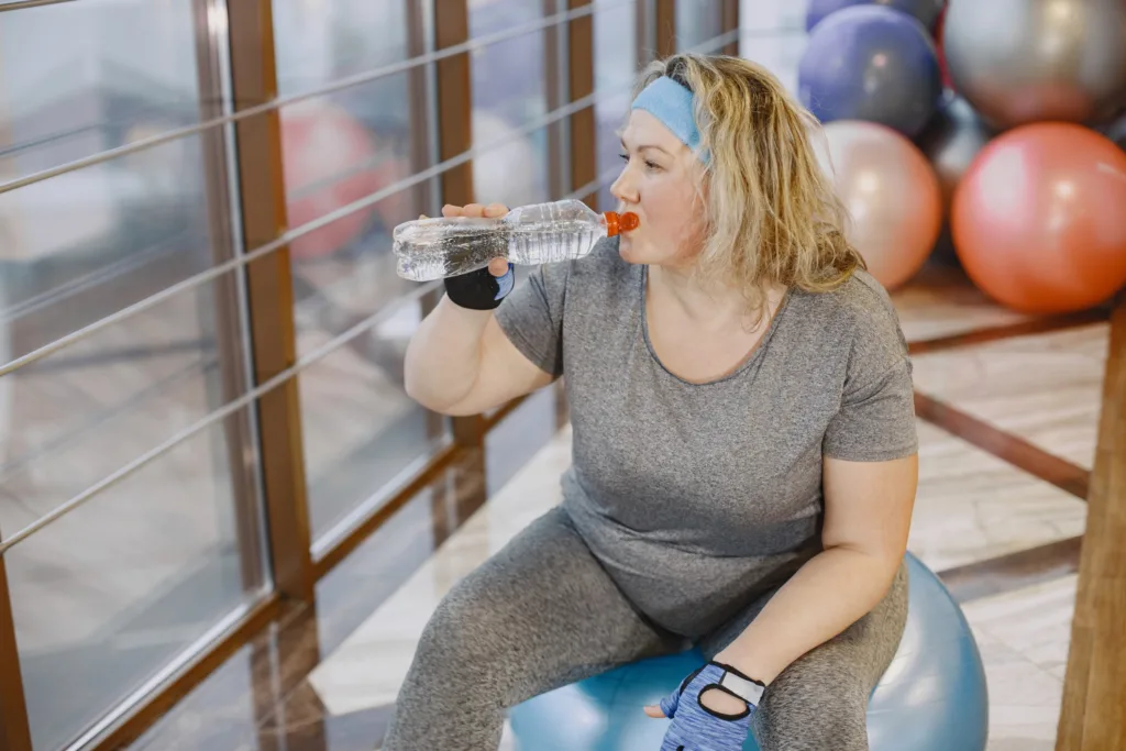 uma senhora na academia bebendo agua, sentada numa bola de fisioterapia.