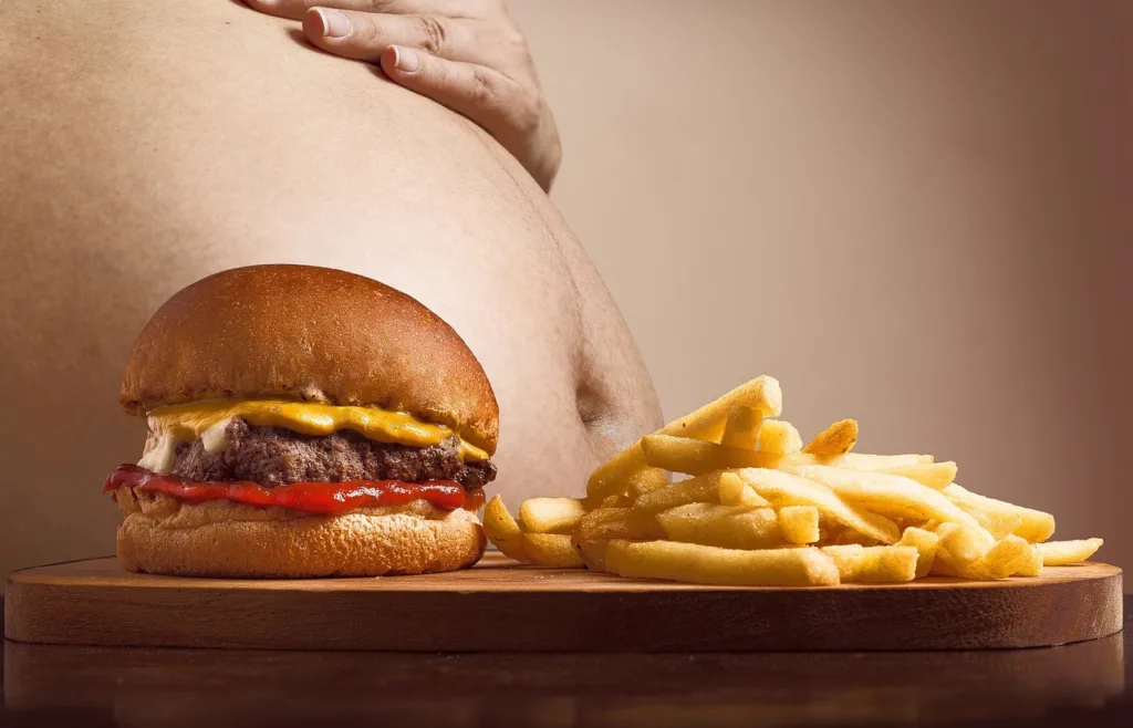 Hamburguer e batata fritas, alimentos gordurosos, numa mesa, com a barriga de uma pessoa obesa.