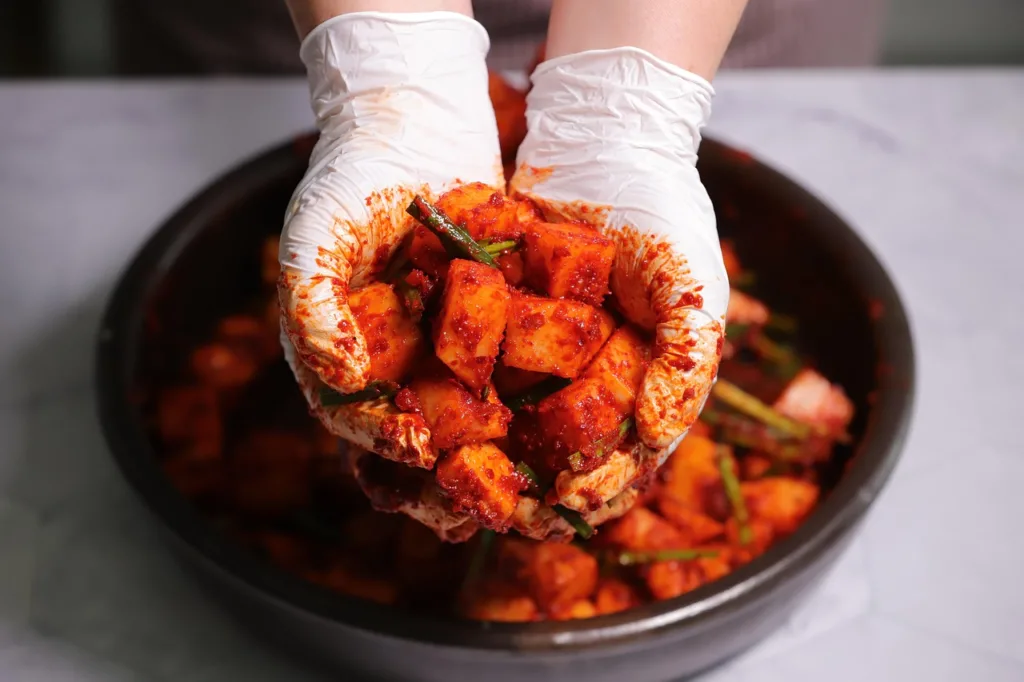uma mão com luvas, segurando uma amostra de kimchi em um prato.