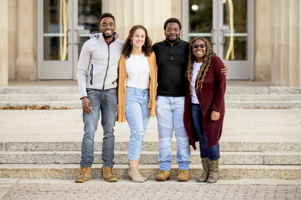 quatro jovens, dois casais, abraçados em linha, ao ar livre.
