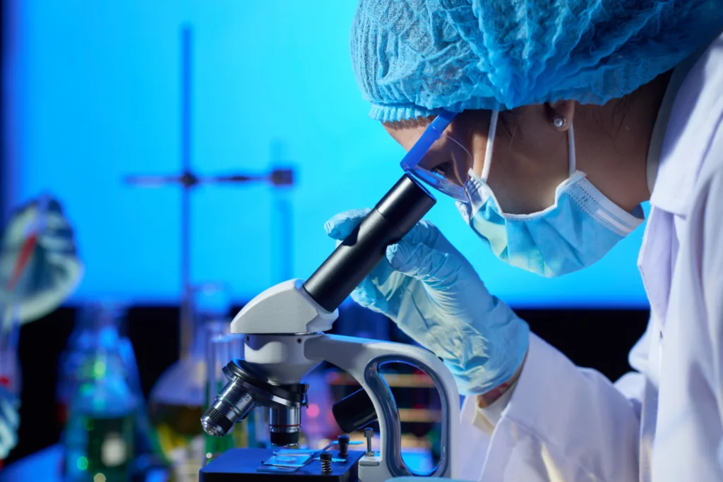 uma cientista com proteção de mascara, observando bactérias através de um microscópio.
