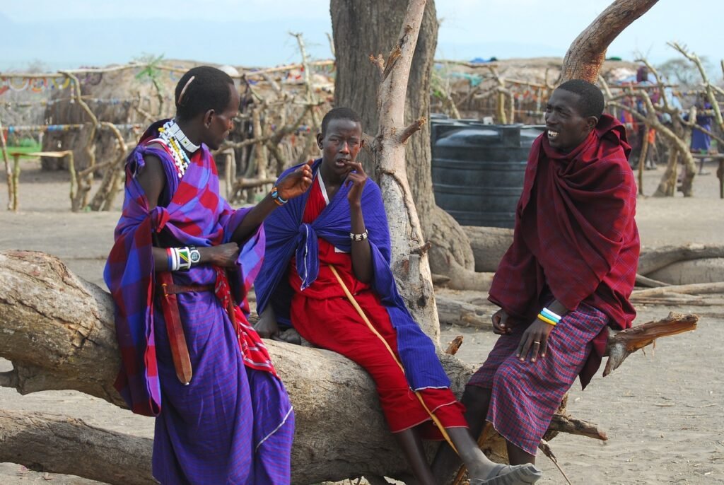 3 homens da tribo maasai sentados em um tronco de arvore, conversando.