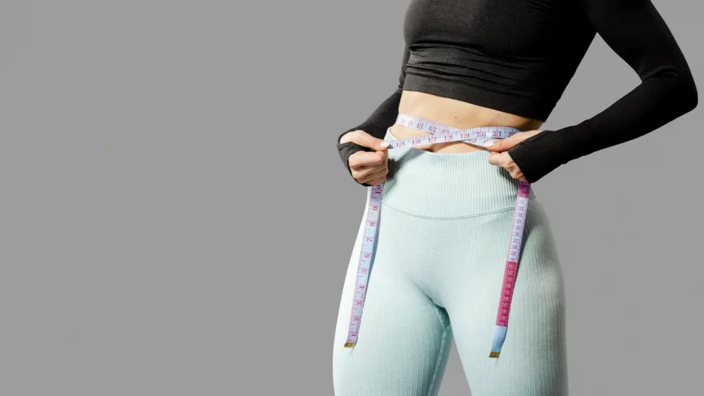 foto do corpo de uma jovem em trajes de ginastica medindo sua cintura com uma fita métrica.
